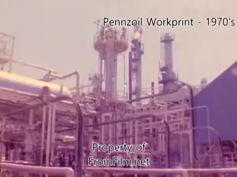 The still from 'Pennzoil Factory/Refinery Workprint - 1980's' showcases an industrial scene typical of a refinery. It features complex machinery, tall distillation towers, and numerous piping structures, all under a slightly pinkish hue typical of 16mm film. The image reflects the operational aspects of oil refining during that era. Notably, there are indications of debris within the film gate, which is often a characteristic of older film stock. The accompanying music is from Underbelly, enhancing the film's ambiance.