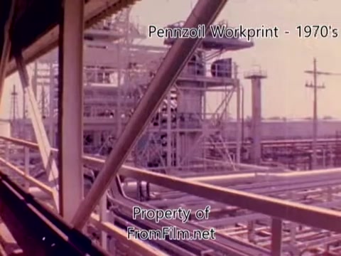 The still from the 'Pennzoil Factory/Refinery Workprint - 1980s' captures a industrial scene showcasing the refinery's extensive piping infrastructure and equipment. The vibrant colors suggest a 1970s aesthetic, typical for film from that era. There's visible debris in the film gate, indicating the film's age and condition. Additionally, the background music features tracks by Underbelly, enhancing the visual experience with atmospheric sounds.