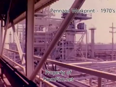The still appears to depict a scene from a Pennzoil refinery workprint from the 1970s, showcasing industrial structures typical of oil refinery operations. The image features scaffolding, pipelines, and large equipment, reflecting the busy atmosphere of a manufacturing environment. The colors seem somewhat faded, suggesting wear typical in older film formats like 16mm. Additionally, the mention of debris in the film gate implies visual artifacts that are characteristic of film projection, not the film itself. The accompanying music tracks by Underbelly add a dramatic auditory backdrop to the visual content.