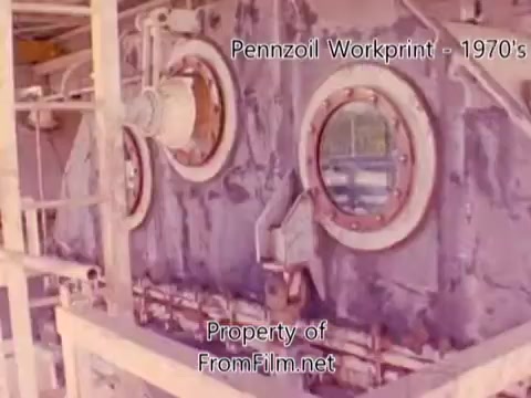 The still from the 'Pennzoil Factory/Refinery Workprint - 1980's' showcases industrial machinery, featuring circular porthole windows embedded in a textured surface, likely part of the refinery's structure. The coloration reflects the era's aesthetic, with visible wear and tear indicating age. This scene captures the essence of refinery operations, evoking a sense of industrial history. The film is accompanied by tracks from Underbelly, enhancing the viewing experience.