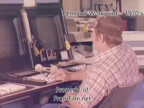 The still features a man in a cap, sitting at a desk with multiple monitors, likely engaged in editing or processing footage related to the Pennzoil factory or refinery. The setting appears to reflect a 1970s or 1980s industrial environment, emphasizing a vintage aesthetic through the film's quality and color. It includes some visible debris in the film gate, typical of older film prints. The accompanying music by Underbelly enhances the nostalgic vibe, adding atmosphere to the workprint.