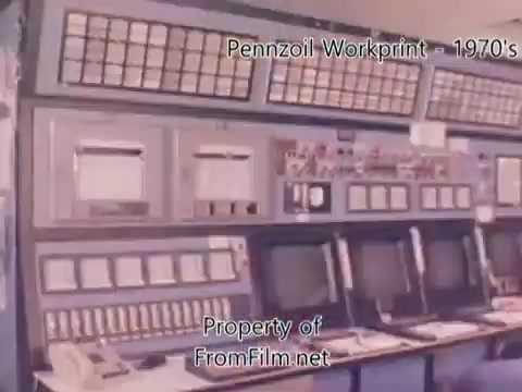 The still captures a control room from the Pennzoil refinery, showcasing an array of gauges, buttons, and monitors typical of the 1970s industrial design. The equipment appears worn, with some visible debris in the film gate, suggesting it’s a workprint from a 16mm negative. The overall atmosphere reflects the operational environment of a petroleum refinery during that era. The accompanying music from Underbelly adds a contemporary auditory layer to the vintage visuals.