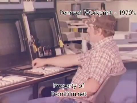 The still depicts a worker seated at a control panel in the Pennzoil refinery, showcasing a retro environment typical of the 1980s. The worker, wearing a plaid shirt and a cap, is engaged with equipment featuring buttons and screens. The film quality suggests it comes from a 16mm negative, with noticeable debris in the film gate, adding a characteristic vintage look. The accompanying music tracks from Underbelly enhance the atmosphere of the work environment.