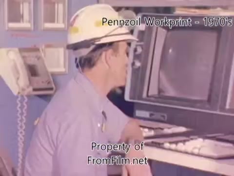 The still from the 'Pennzoil Factory⧸Refinery Workprint - 1980s' features a man wearing a hard hat, seated in front of equipment that includes a monitor and control panel. The background shows industrial elements, typical of a refinery setting. The film exhibits some debris in the film gate, indicative of age or wear. The music playing is by Underbelly, featuring tracks such as 'I Hear Voices,' 'Asterism,' 'Italy Jr.,' and 'Land's End,” enhancing the atmosphere of the scene.