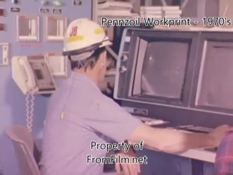 The still from 'Pennzoil Factory⧸Refinery Workprint - 1980's' depicts a worker seated in front of a control panel, wearing a hard hat labeled 'JB.' He appears focused on monitoring screens, suggesting oversight of refinery operations. The setting is likely a control room, characterized by various instruments and gauges. The overall aesthetic hints at a vintage look typical of the 1970s or 1980s industrial environments, with a slightly muted color palette that captures the period's filmic quality. Debris in the film gate adds an authentic touch of age to the footage.