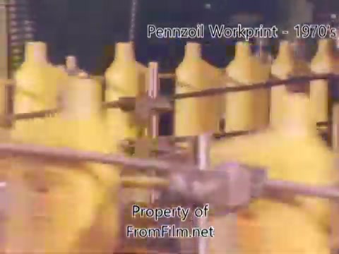 The still from the 'Pennzoil Factory/Refinery Workprint - 1980s' shows a dynamic scene in a refinery setting. It features an assembly line with yellow oil containers moving steadily. The film gate has some debris visible, indicating it's a workprint from a 16mm negative. The accompanying music by Underbelly adds a modern auditory backdrop to the industrial visuals.