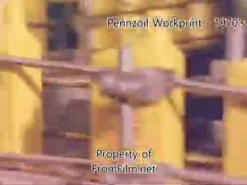 The still from the 'Pennzoil Factory/Refinery Workprint - 1980s' features industrial scenes within a refinery, characterized by yellow machinery and pipes. The image appears to show the inner workings of the plant, with some visible debris in the film gate, indicating its age. The music accompanying the visuals is from Underbelly, featuring a mix of tracks that enhance the atmosphere of the footage. Overall, it presents a nostalgic glimpse into refinery operations during that era.