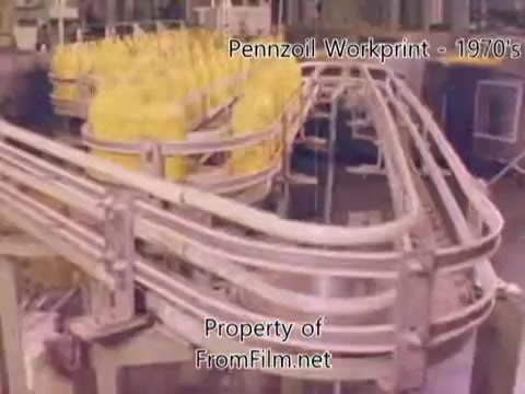 The still captures a scene from the Pennzoil factory, showcasing a production line filled with yellow containers. The machinery features intricate conveyor belts, highlighting the industrial process of oil packaging. This segment is part of a workprint from the 1970s, which reflects the era's manufacturing practices. Notably, there's visible debris in the film gate, indicating wear or age of the 16mm negative. The accompanying music tracks from Underbelly, such as 'I Hear Voices' and 'Asterism,' add an atmospheric backdrop to the imagery.
