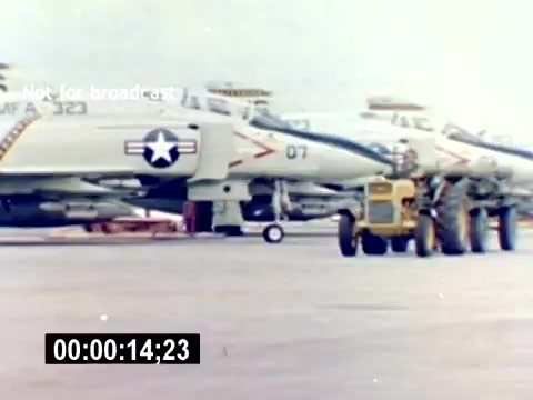 The still shows two military aircraft, likely F-4 Phantoms, on a tarmac with a ground vehicle in the foreground. Personnel in military attire are working near the aircraft, engaging in loading or maintenance activities. The setting appears to be a military base focused on aviation operations. The absence of sound suggests it’s part of a silent film documenting USMC aviation ordnance and recruitment efforts.