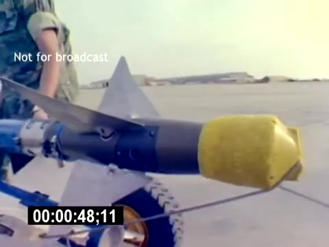 The still from the 'USMC Aviation Ordnance Recruiting Film - Silent' features a close-up view of a military ordnance device, likely an explosive or missile component, which is mounted on a transport dolly. The device has a distinct yellow nose cone, indicating its operational purpose. A service member dressed in military camouflage is positioned nearby, emphasizing the practical aspect of ordnance handling. The background suggests a military airfield setting, reinforcing the film's focus on U.S. Marine Corps aviation ordnance recruitment. A timestamp and the note 'Not for broadcast' are also displayed on the image.