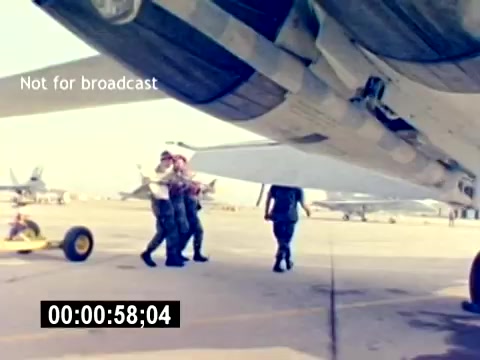 The still from the 'USMC Aviation Ordnance Recruiting Film - Silent' showcases a military setting on an airfield. It features personnel moving beneath the wing of a military aircraft, likely engaging in some form of ordnance or maintenance operation. In the background, additional military planes can be seen, emphasizing the aviation theme. The scene appears focused on teamwork and the operational environment of the U.S. Marine Corps. The timecode at the bottom indicates it is part of a larger sequence.