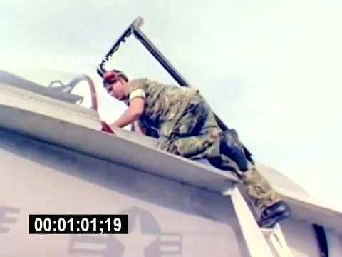 The still captures a member of the US Marine Corps climbing onto the wing of an aircraft. Dressed in military fatigues, the individual demonstrates maintenance or loading activities on the aircraft, highlighting the hands-on nature of aviation ordnance work. The background features a clear sky, emphasizing the operational environment of military aviation. The absence of sound suggests a focus on visual storytelling.