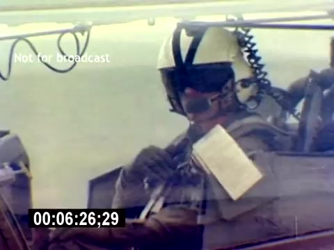 The still features a pilot inside a cockpit, wearing a flight helmet and headset, preparing for a mission. The focus is on the pilot's serious demeanor as they handle equipment, indicative of military aviation operations. The image is marked 'Not for broadcast' and includes a timecode at the bottom, suggesting it is part of a training or recruitment film for USMC Aviation Ordnance.