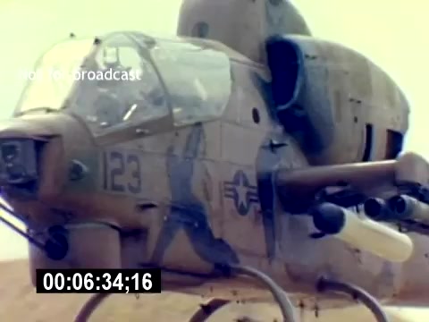 The still features a close-up view of a military helicopter, likely an AH-1 Cobra, showcasing its detailed cockpit and weaponry. The vehicle is prominently marked with the number '123' and military insignia, set against a beige landscape that suggests a training or operational environment. The focus highlights the aircraft's design and armament, reflecting its role in military aviation.