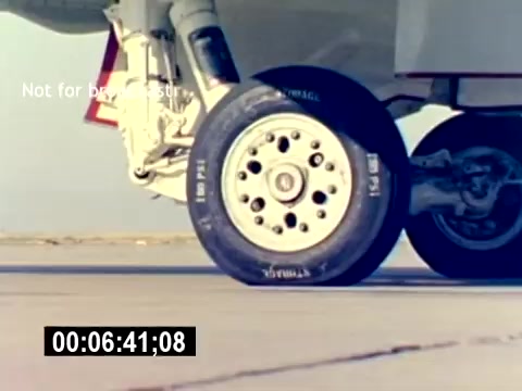 The 16mm still shows a close-up of an aircraft's landing gear, focusing on the wheel and tire. The scene likely captures the aircraft preparing for takeoff or landing on a runway, emphasizing the mechanics of aviation ordnance. The color and clarity suggest a vintage aesthetic typical of military recruitment films. A timestamp is visible in the corner, indicating the specific moment in the film.