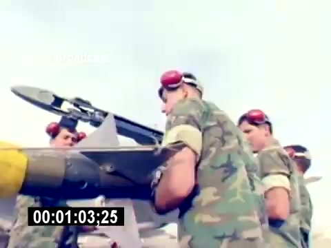 The still from 'USMC Aviation Ordnance Recruiting Film - Silent' depicts a group of military personnel in camouflage uniforms and red ear protection as they handle ordnance, specifically a missile or bomb, on a tarmac. The focus is on teamwork and precision, showcasing the process of preparing aviation ordnance. The scene emphasizes the technical and operational aspects of military aviation duties.