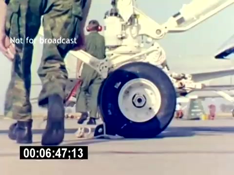 The still from the 'USMC Aviation Ordnance Recruiting Film - Silent' captures a close-up view of an aircraft's wheel and landing gear on a runway. In the foreground, a Marine in camouflage uniform walks past, suggesting a focus on the operational environment of military aviation. The scene emphasizes the technical aspects of aviation ordnance preparation, underlining the professionalism and activity involved in the Marines' aviation sector. The image’s composition centers on the equipment, signaling the importance of logistics and maintenance in military operations.