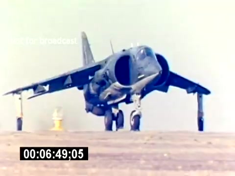The still depicts a military aircraft, likely a Harrier jet, taxiing on a runway. The setting is presumably a military base, characterized by a barren landscape and a control tower or marker in the background. The image captures the aircraft's powerful design and operational readiness, highlighting its role in aviation ordnance for the US Marine Corps. The timestamp indicates the specific moment within the film.