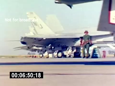 The still from the 'USMC Aviation Ordnance Recruiting Film - Silent' depicts a U.S. Marine in green flight gear walking near a military aircraft on the tarmac. The aircraft, likely a fighter jet, is visible in the background with visible ordnance. The image conveys a sense of military operations and the role of ordnance specialists in aviation. The scene is bright and showcases a clear day, emphasizing the operational environment.