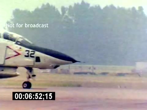 The still shows a military aircraft, specifically a fighter jet, taxiing on a runway. The plane is marked with the number '32' and military insignia. The background appears slightly blurred, giving a sense of motion, and there are hints of an airfield environment. A timestamp and a 'Not for broadcast' watermark are visible in the frame, suggesting this is a still from a film not intended for public airing. The overall atmosphere conveys a focused military context related to aviation ordnance.
