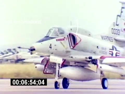 The still shows a military aircraft, likely an AV-8 Harrier or a similar jet, with identifying markings, including a red and blue insignia and the number '4' prominently displayed. The aircraft is on a runway with other planes visible in the background. The scene captures a moment of aviation activity, emphasizing the function and readiness of USMC aviation ordnance. The time code indicates the clip's duration in the film.