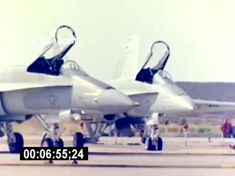 The still features two military aircraft parked side by side on an airfield. Both planes have their cockpit canopies open, suggesting preparation for operation or maintenance. The background shows a clear sky and possibly mountainous terrain, highlighting a military setting. The focus appears to be on showcasing the aircraft, which likely represent the capabilities of USMC aviation ordnance.