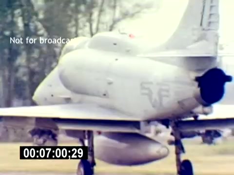 The 16mm still features a military aircraft, likely a Marine Corps fighter jet, in a display setup. The aircraft is shown from a side angle, revealing its sleek design and undercarriage, which houses ordnance. The scene is outdoors, surrounded by greenery, indicating a military airfield. The text 'Not for broadcast' suggests it is intended for private or training use. The timestamp at the bottom signifies the film's time code.