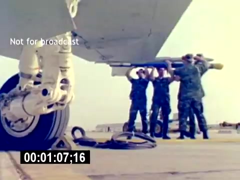 The still captures a group of Marines in military fatigues working together to load a missile onto an aircraft. The focus is on teamwork and the technical aspect of ordnance handling. The framing emphasizes the wheel of the aircraft and the men positioned under the wing, highlighting their coordinated effort. The environment appears to be a military airfield, indicative of aviation operations. The absence of sound suggests a focus on visual training.
