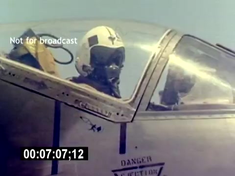 The still features a close-up of a pilot in a cockpit, wearing a helmet and oxygen mask. Another individual, presumably a crew member, is visible in the background. The aircraft has a metallic surface, showcasing operational details. The image captures a moment in a military aviation context, emphasizing readiness and the environment of the USMC.