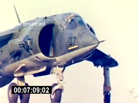 The still shows a close-up of a military aircraft, likely a helicopter or VTOL, identified by the '07' marking on its fuselage. The image captures the aircraft in a stationary position, highlighting its design and features, particularly the landing gear and cockpit area. The background appears light, suggesting a clear sky or open environment, typical of a military training or demonstration setting. The absence of sound indicates that this is a silent film intended for visual impact.