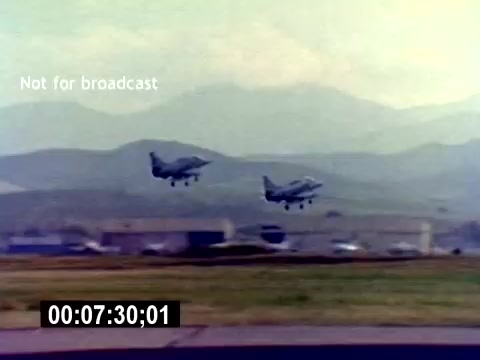 The still depicts two military aircraft taking off or landing in a mountainous landscape. The background features an airbase with buildings and vehicles, suggesting an active military environment. The scene highlights the operation of USMC aviation, likely emphasizing the capabilities and excitement of military aviation to attract recruits. The timestamp indicates it's from 7 minutes and 30 seconds into the film.