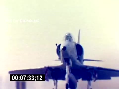 The still depicts a military aircraft, likely a jet, taking off or in a commanding position. The image is characterized by its vintage 16mm quality, featuring a slightly blurred background that highlights the aircraft. The context suggests this is part of a U.S. Marine Corps film focused on aviation ordnance recruiting, aiming to showcase the aircraft's capabilities and the role of ordnance specialists. The absence of sound emphasizes the visuals, which convey a sense of power and precision in military aviation.