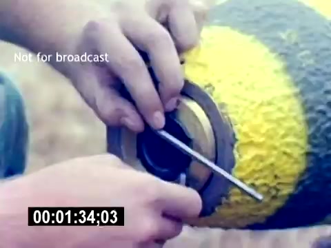 The still from the 'USMC Aviation Ordnance Recruiting Film' depicts a close-up of a person’s hands working on a yellow and green munition. The individual is using a metal tool, possibly to secure or adjust a component of the ordnance. The focus on the hands and the ordnance suggests an emphasis on precision and technical skill in aviation ordnance handling. The image is time-stamped at 01:34:03 and marked as not for broadcast.