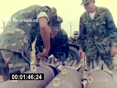 The still from the 'USMC Aviation Ordnance Recruiting Film - Silent' shows three Marines in camouflage uniforms working together around several large bombs or ordnance on the ground. The focus is on their hands as they appear to be inspecting or handling the equipment. The scene conveys a sense of teamwork and technical expertise in a military setting. The visible timecode indicates it's part of a longer film.