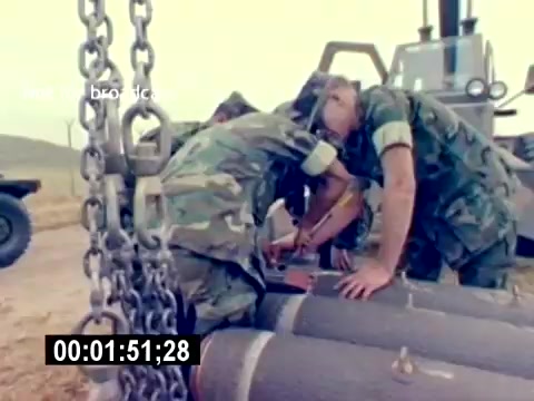 This 16mm still from the 'USMC Aviation Ordnance Recruiting Film - Silent' depicts a scene where two military personnel are working closely together on munitions. They appear to be focused on preparing or handling ordnance, with visible equipment and tools nearby. The setting suggests an outdoor environment, likely on a training field. Their camouflage uniforms indicate their military affiliation, emphasizing a hands-on approach to aviation ordnance work.