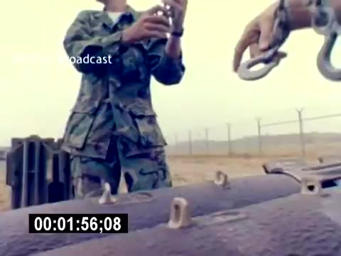 The still from the 'USMC Aviation Ordnance Recruiting Film - Silent' showcases a military setting, with personnel in camouflage uniforms handling equipment. One individual is holding an object, possibly a bomb or ordnance, while another is using metal hooks, likely for securing or deploying the equipment. The background features an outdoor area with a wire fence, suggesting a base or training ground. The overall focus is on the technical aspects of aviation ordnance.