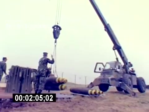 The still shows a military setting where personnel are involved in handling ordnance. A crane is lifting large munitions, while several uniformed individuals oversee the operation. The environment appears foggy or overcast, contributing to a somber and serious atmosphere. The focus on ordnance handling emphasizes the technical and operational aspects of USMC aviation ordnance duties.
