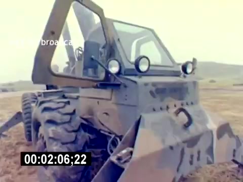 The still from the 'USMC Aviation Ordnance Recruiting Film - Silent' depicts a military vehicle, likely designed for ordnance handling or support. It features a camouflaged exterior, large tires for rugged terrain, and equipment for operational tasks. The vehicle is situated on open land, indicating its use in a field environment. The overall design suggests functionality and adaptability for military operations.