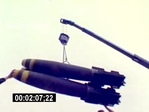 The still depicts a crane lifting two large bombs, characterized by their cylindrical shape and distinct yellow stripes. The scene suggests a military environment, likely involving aviation ordnance operations. The timecode at the bottom indicates the film's progress, and there are no accompanying sounds in this silent film segment. The overall focus is on the logistical aspects of handling munitions.