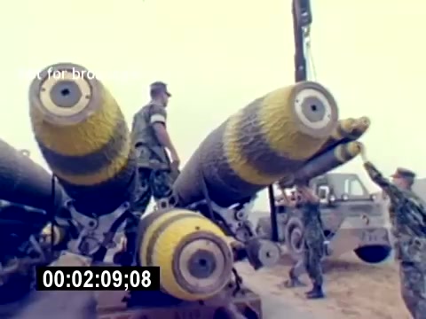 The still depicts U.S. Marines engaged in handling large ordnance, likely bombs, used in aviation. The munitions are painted with distinctive yellow and black stripes, indicating their type or purpose. Marines are seen working together, showcasing teamwork in a military setting, with equipment in the background. The environment appears to be an outdoor area, possibly a loading or operational facility. The lack of a soundtrack suggests a focus on visual elements rather than audio commentary.