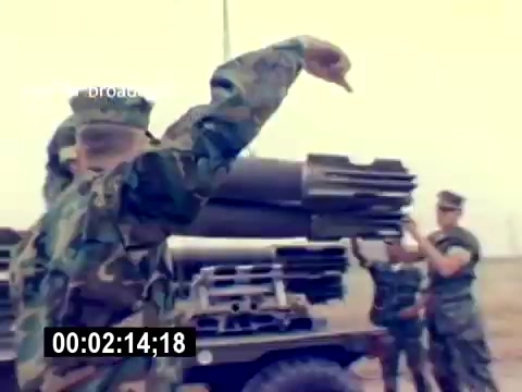 The still from the 'USMC Aviation Ordnance Recruiting Film - Silent' depicts a group of Marines interacting around an artillery piece or missile system. One Marine is gesturing, likely giving directions or demonstrating a procedure, while others appear to be engaged in tasks related to the equipment. The setting suggests a military training environment, emphasizing teamwork and operational readiness. The overall tone is determined and focused on ordnance handling in a tactical context.