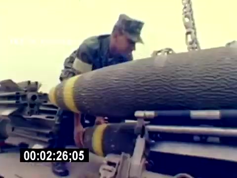 The still features a U.S. Marine in uniform, engaged in handling a large ordnance item, likely a bomb or missile. The Marine is focused on safely positioning the ordnance, which is cylindrical with yellow markings. The background suggests an operational military environment, possibly on a loading dock or aircraft carrier, reflecting the technical and precise nature of aviation ordnance work. The image emphasizes the hands-on responsibilities of Marines in aviation support roles.