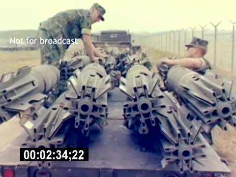 The still captures two U.S. Marines interacting with munitions on a military vehicle. They are surrounded by various ordnance, which appears to be missiles or bombs, displayed neatly in rows. The setting has a military ambiance, with a perimeter fence in the background, suggesting a base or secure area. The equipment emphasizes the focus on aviation ordnance and training. The timecode indicates the specific moment in the film, marking it for reference.