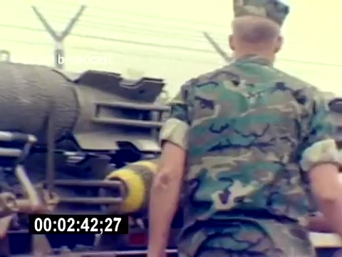 The still from 'USMC Aviation Ordnance Recruiting Film - Silent' shows a service member in a camouflage uniform walking towards a military vehicle that appears to be equipped for ordnance handling. The background features a security fence and part of the vehicle, emphasizing a focus on military operations and training. The timecode at the bottom indicates the specific moment within the film.