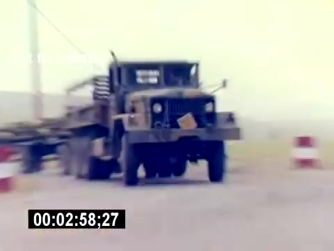 The still from the 'USMC Aviation Ordnance Recruiting Film - Silent' depicts a military truck, possibly an ordnance transport vehicle, moving along a road marked by barriers. The environment appears dusty or foggy, suggesting a training or operational area. The image captures a moment highlighting the logistics and equipment involved in Marine Corps aviation ordnance operations. The timecode at the bottom indicates the exact point in the film.