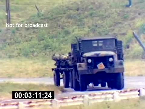 The still depicts a military truck on a roadway, transporting ordnance. The truck is camouflaged, suggesting a military context, and is likely part of a training or demonstration scenario related to USMC aviation ordnance. The grassy terrain in the background indicates an outdoor setting, possibly a military base or training area. The timestamp suggests it is a moment captured during a longer film segment focused on recruitment or operational procedures.