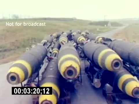 The still from the 'USMC Aviation Ordnance Recruiting Film - Silent' shows a close-up view of several bombs or ordnance items lined up on a transportation vehicle or flatbed. The bombs are cylindrical with distinctive yellow and black markings. The background features a grassy area with a slightly overcast sky, suggesting a military or training environment. The time code at the bottom indicates the specific moment in the film. The caption 'Not for broadcast' implies this is a restricted or behind-the-scenes shot.