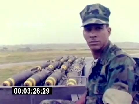 The still features a young Marine in uniform, sitting in a truck or vehicle. The background shows a military airfield with ordnance, including missiles or bombs, neatly arranged. The atmosphere appears overcast, suggesting a utilitarian military setting. The Marine's expression is serious, conveying focus and readiness, indicative of the recruitment theme centered on aviation ordnance roles.