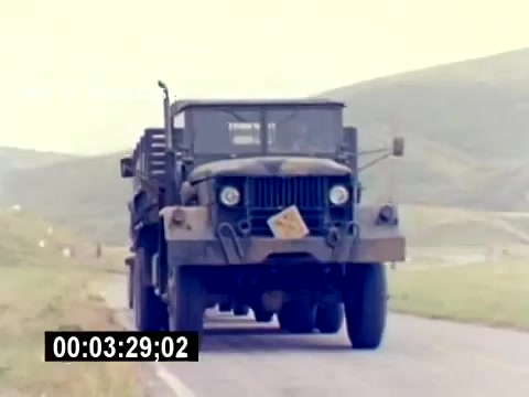 The still depicts a military truck moving along a rural road, set against a backdrop of rolling hills. The vehicle appears rugged and is likely equipped for transporting ordnance or personnel. The visible license plate indicates military use. The scene conveys a sense of operation and movement, typical for a recruiting film aimed at showcasing USMC aviation ordnance capabilities.