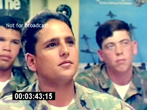 The still features a group of young Marines, likely in a classroom or briefing setting. The focus is on one Marine in the foreground, who appears attentive and engaged. The others in the background also look focused, indicating that they are receiving instruction or information related to aviation ordnance. Various military-themed posters are visible on the walls, providing context to their training environment. The image captures a moment of camaraderie and concentration among the servicemen.